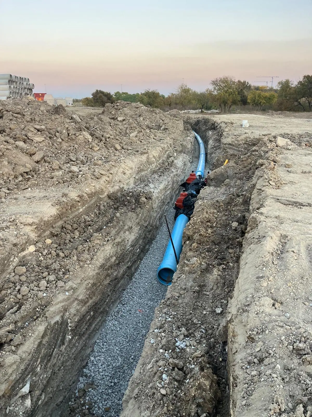 Water line with red gate valves installed in a rock-bedded trench at dusk